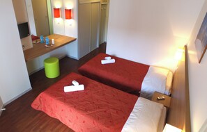 Une chambre d’hôtel avec deux lits, un petit bureau, une poubelle verte et un téléviseur.