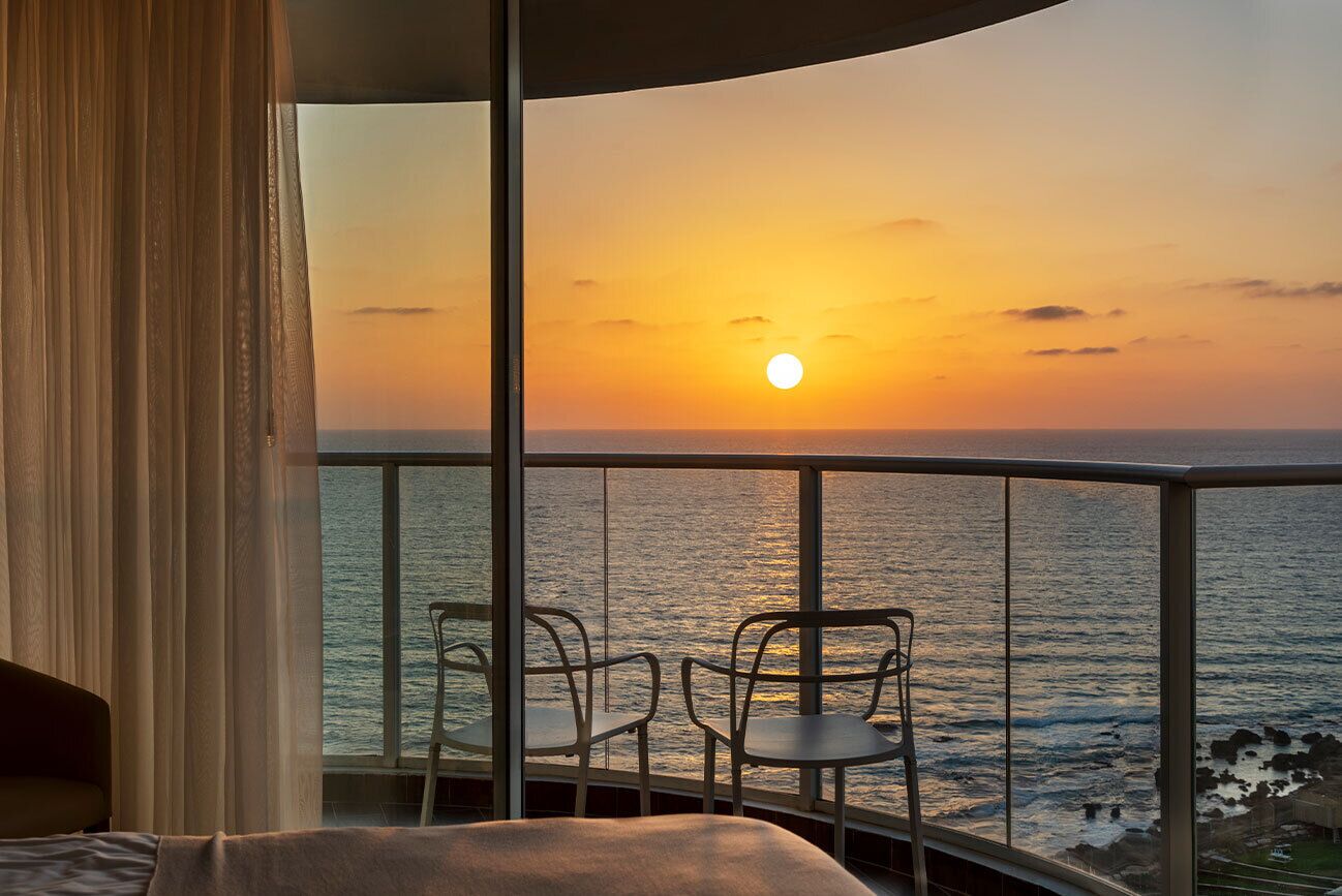 Un balcón con vista al atardecer sobre el océano, una cama y dos sillas.