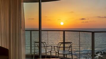 Un balcón con vista al atardecer sobre el océano, una cama y dos sillas.