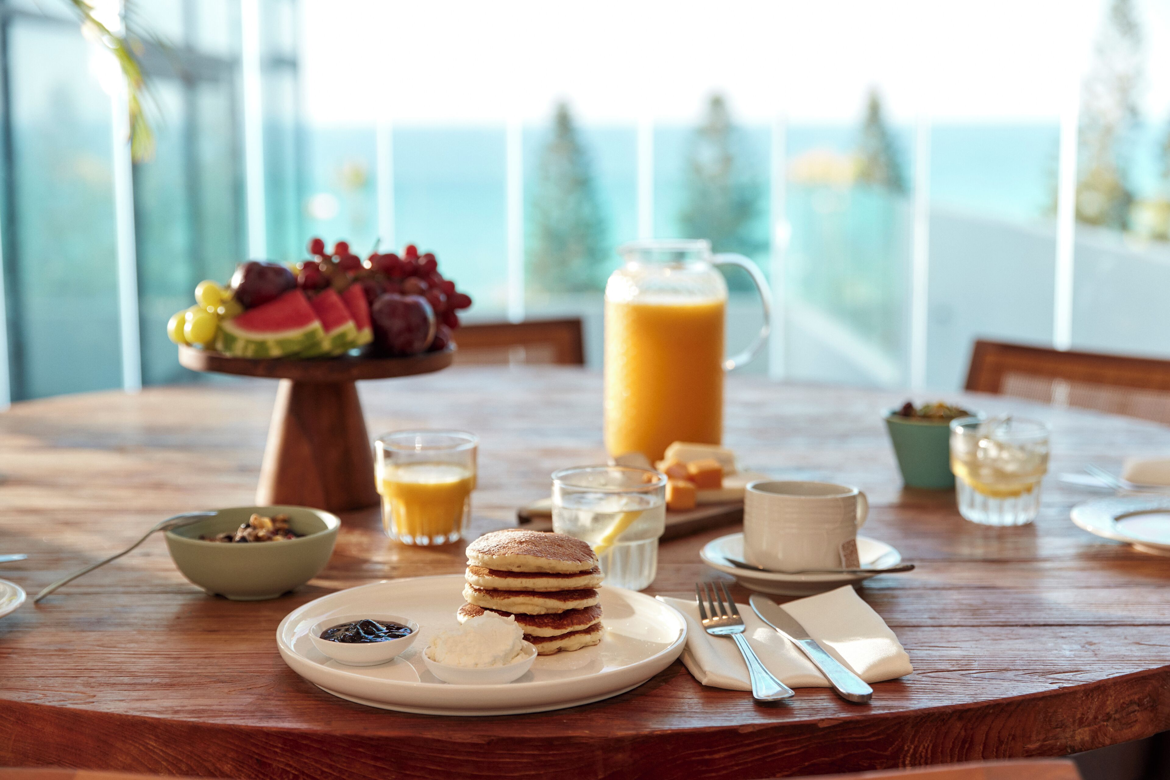 Une table de petit-déjeuner avec des crêpes, une carafe de jus d’orange et un saladier de fruits.