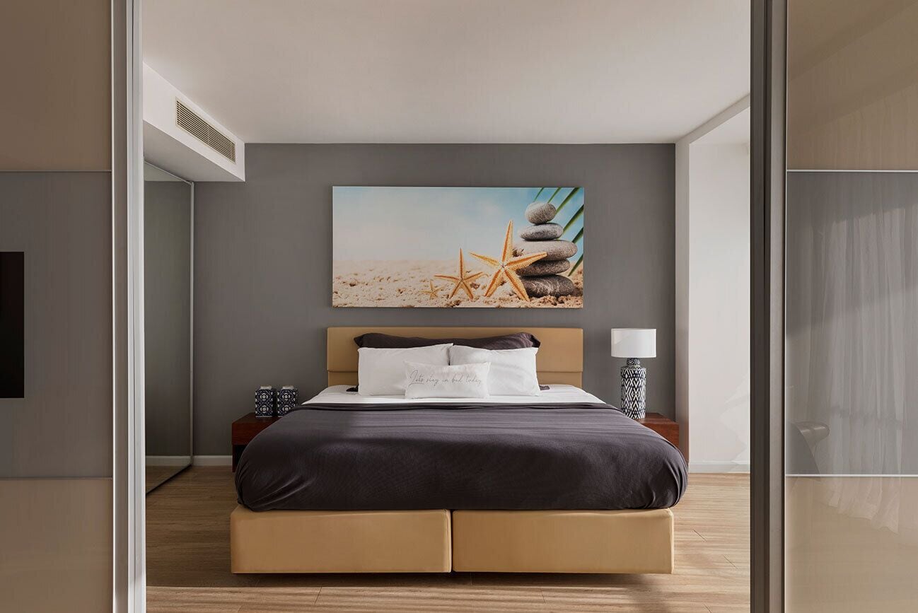 A modern hotel room with a bed, a nightstand, a lamp, and a beach-themed artwork on the wall.