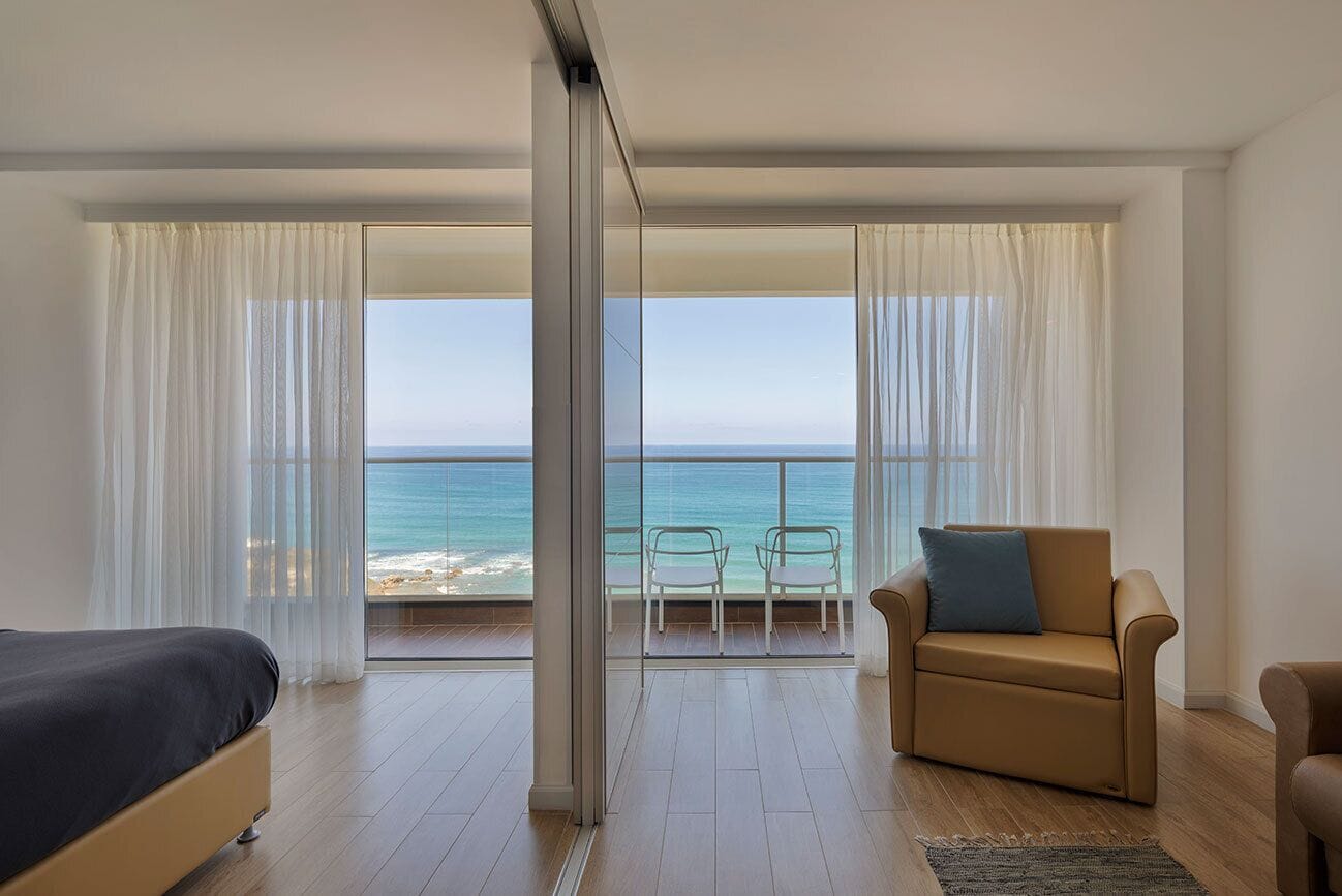 Una habitación con cama, sofá, sillón y vistas al océano a través de amplios ventanales.