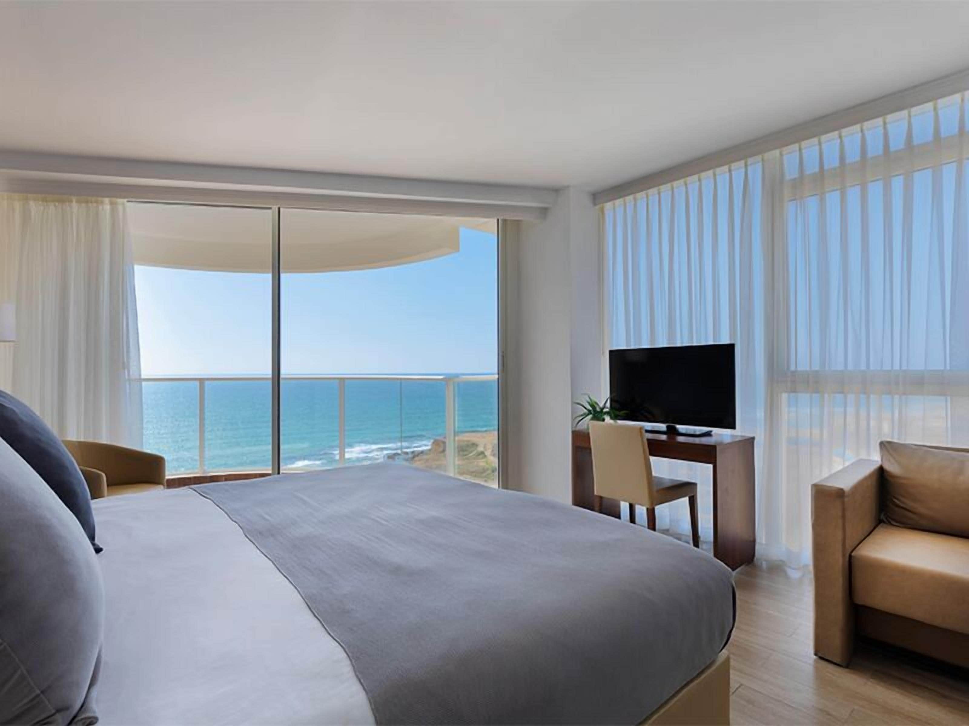Een hotelkamer met een groot bed, een bureau met een televisie en uitzicht op de oceaan.