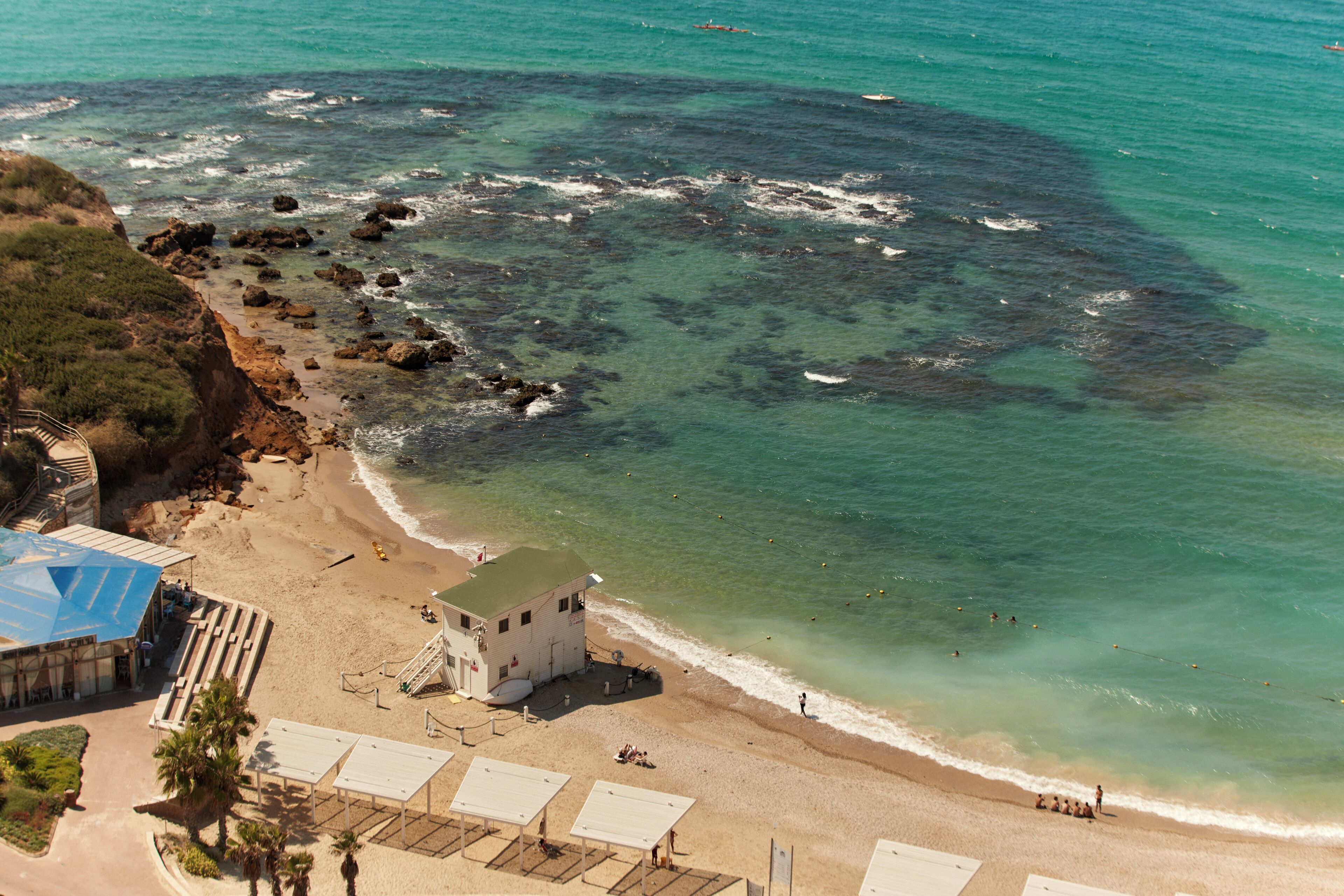 An aerial view of a beach with a building, clear turquoise waters, and rocky areas.