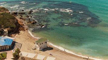 An aerial view of a beach with a building, clear turquoise waters, and rocky areas.