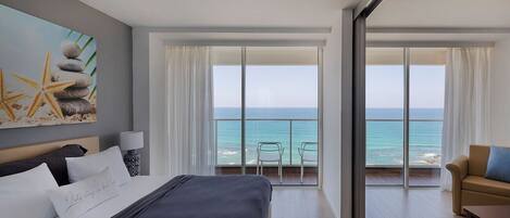 Een hotelkamer met een bed, uitzicht op de oceaan, een balkon met stoelen en een schilderij aan de muur.