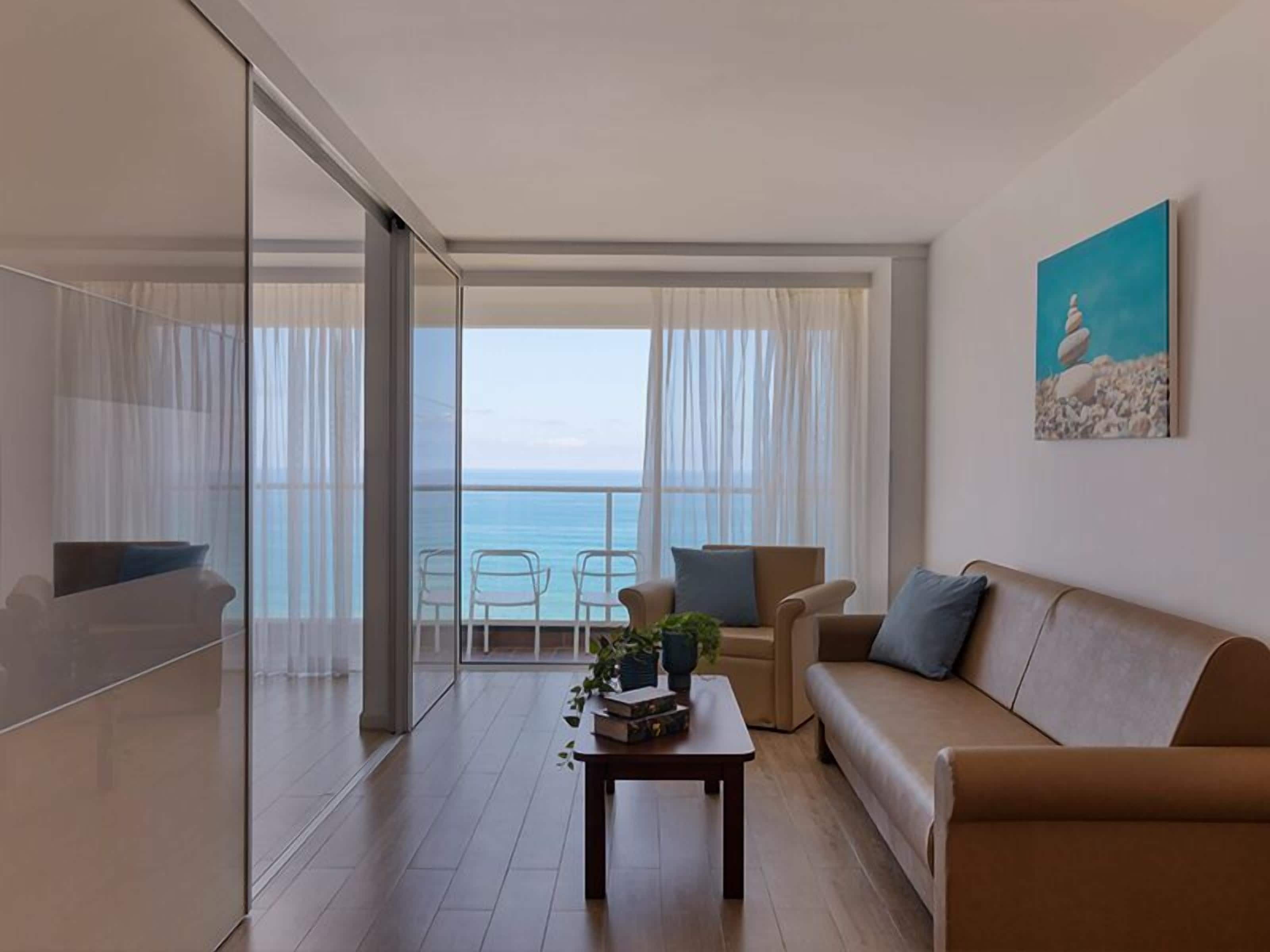 Una sala de estar moderna con un sofá, sillas, una mesita de centro y vistas al mar a través de amplios ventanales.
