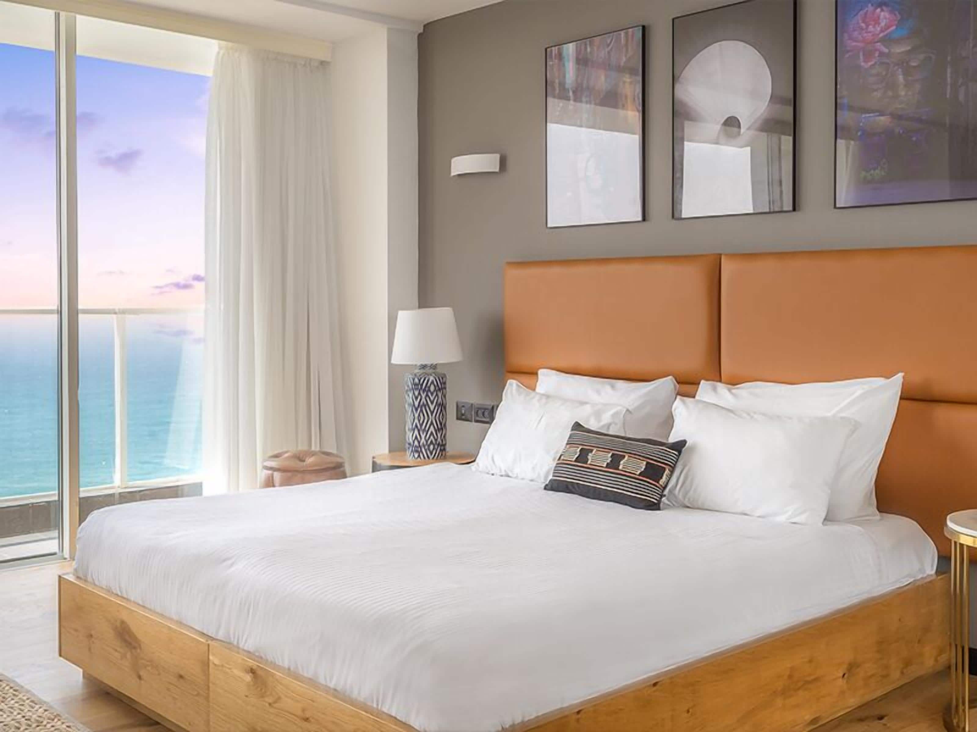 A large bed with white linens, a wooden headboard, and a view of the ocean through a large window.
