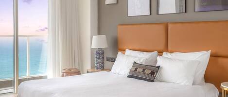 A large bed with white linens, a wooden headboard, and a view of the ocean through a large window.