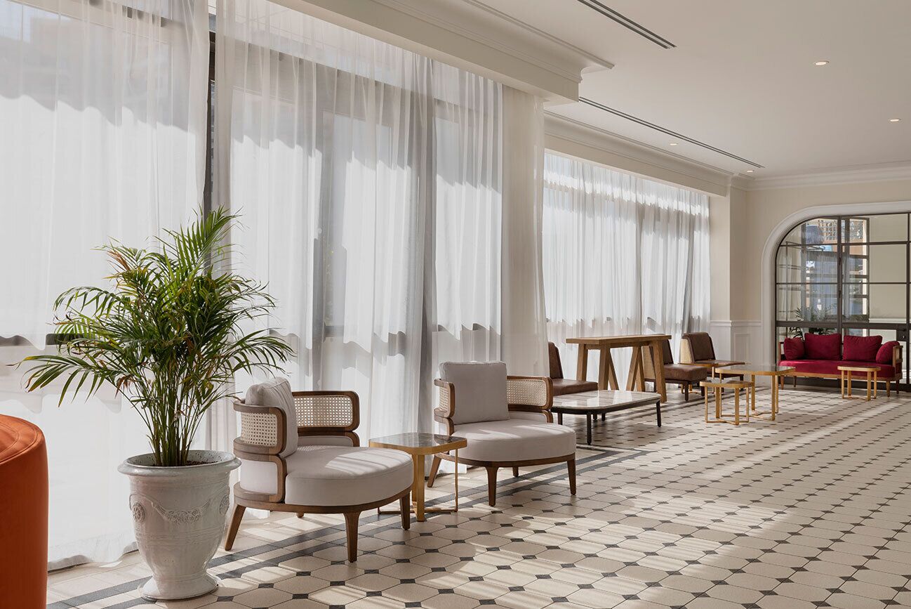 A hotel lobby with a checkered floor, large windows with sheer curtains, a potted plant, and a red sofa.