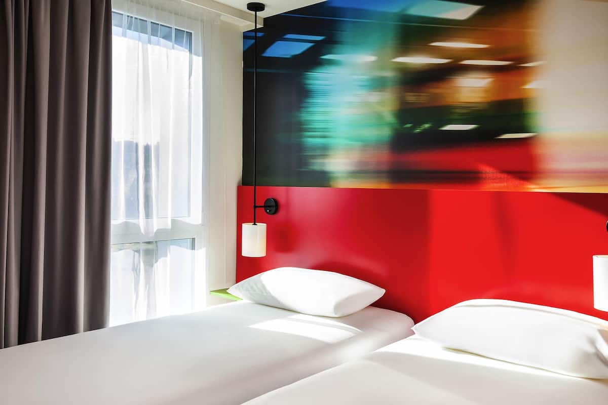 A hotel room with a bed, a window with curtains, and a wall with a red panel.