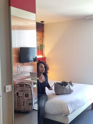 Una habitación de hotel con una cama, televisión en la pared, una maleta y un escritorio con lámpara.