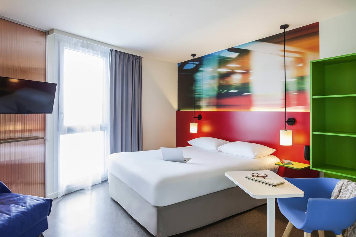 A modern hotel room with a bed, a desk, a chair, a TV, and a green storage unit.