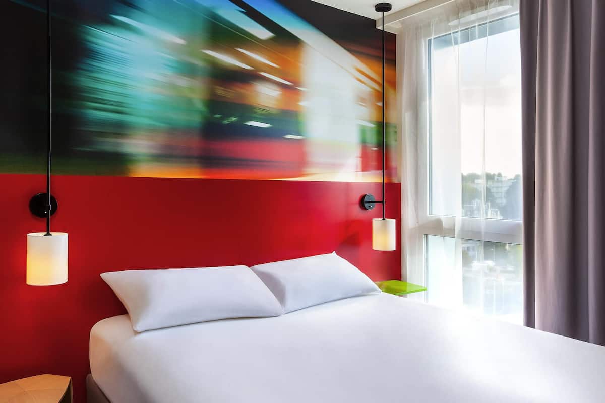 A modern hotel room with a bed, two bedside tables, a large window with curtains, and a colorful abstract painting on the wall.