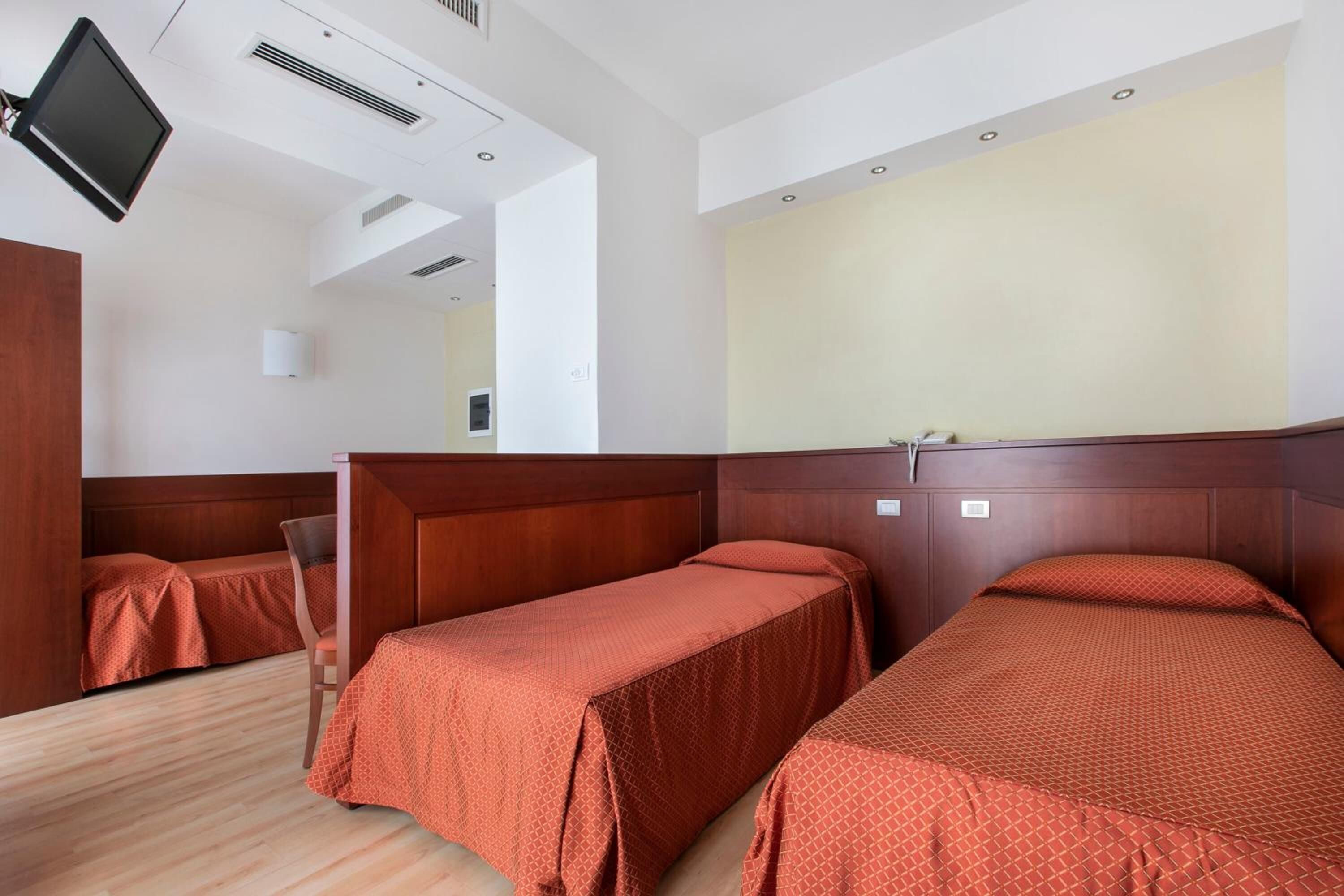 Habitación de hotel con dos camas individuales, cabecero de madera, televisor de pantalla plana montado en la pared y una mesita de noche pequeña.