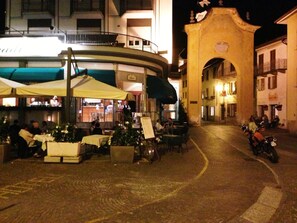 Escena nocturna de una calle con terraza en un restaurante, una motocicleta estacionada al lado y un arco ornamentado al fondo.