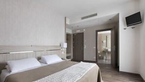 A hotel room with a large bed, a bedside table, a lamp, a TV mounted on the wall, and a bathroom visible through an open door.