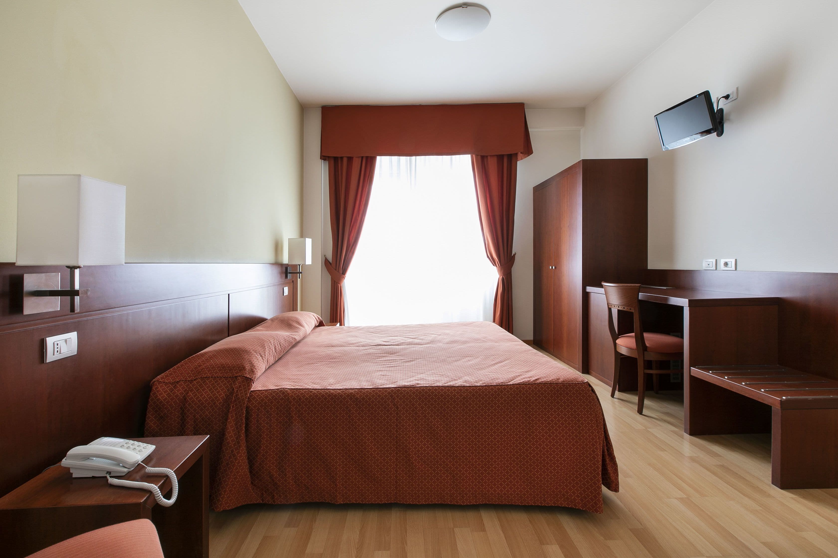 Een hotelkamer met een bed, een bureau, een stoel, een aan de muur bevestigde televisie en een telefoon op het bureau.