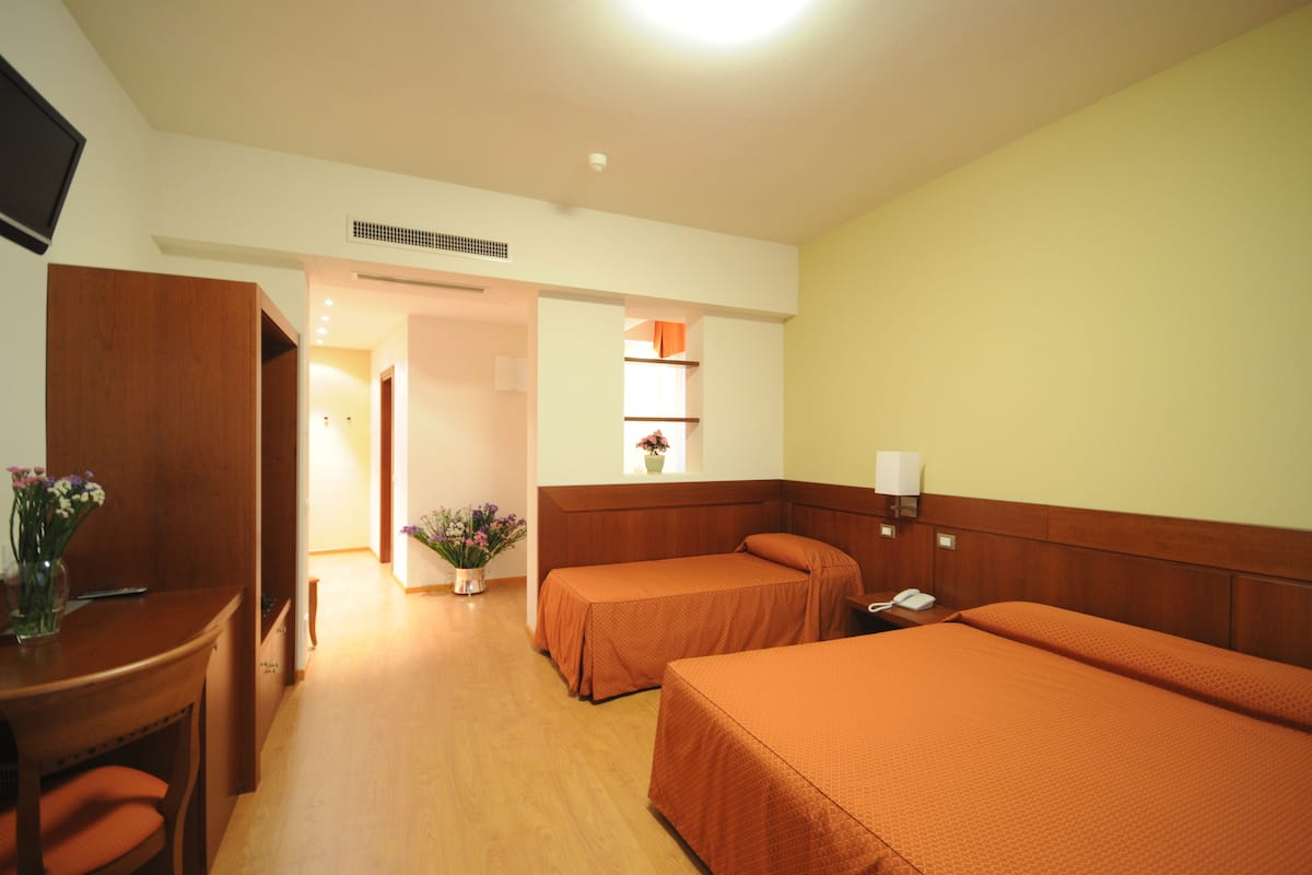 Une chambre d’hôtel avec deux lits, un bureau, une chaise, une petite table ornée d’un vase de fleurs, une lampe et une fenêtre donnant sur un beau paysage.