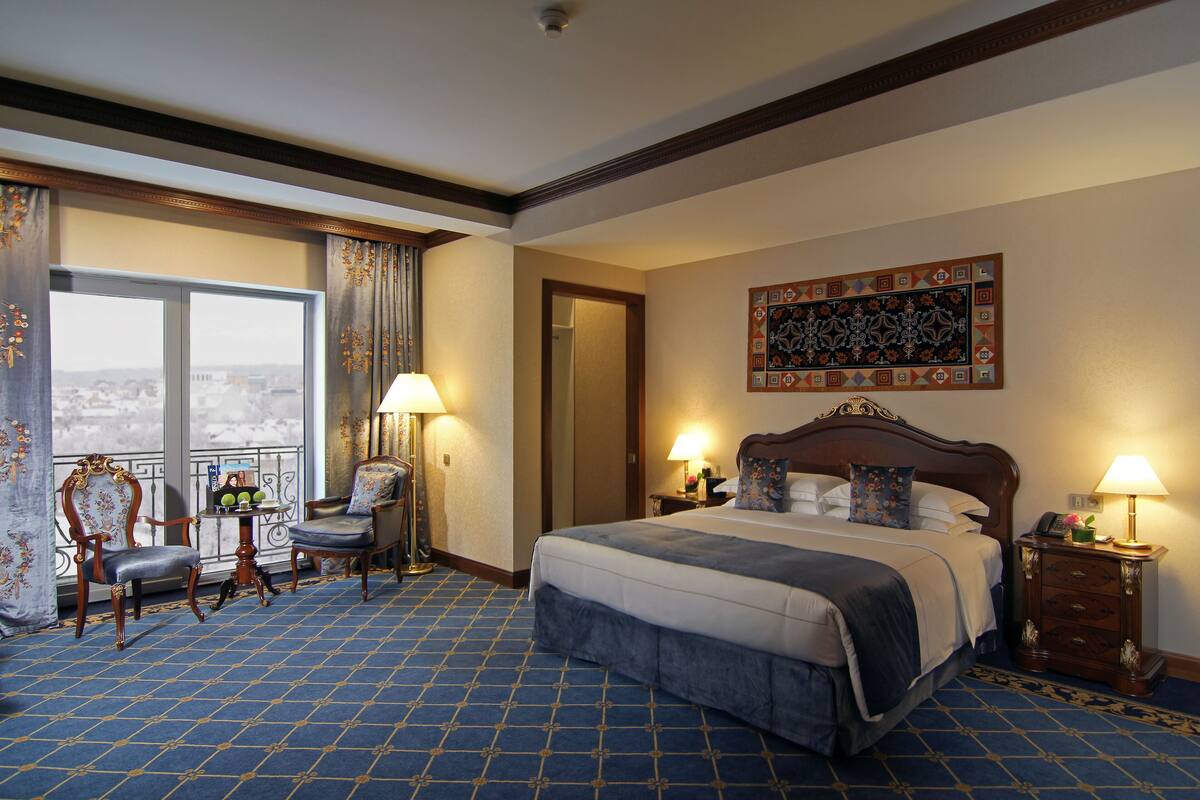 Een hotelkamer met een groot bed, twee stoelen, een klein tafeltje met fruit, een nachtkastje met een lamp en een uitzicht op de stad door een groot raam.