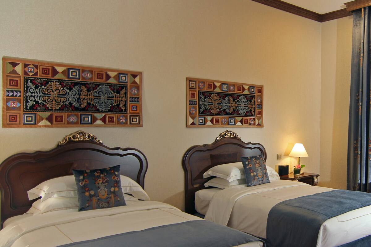 Twee tweepersoonsbedden met hoofdborden, een nachtkastje met een lamp, en twee decoratieve paneeldecoraties aan de muur.