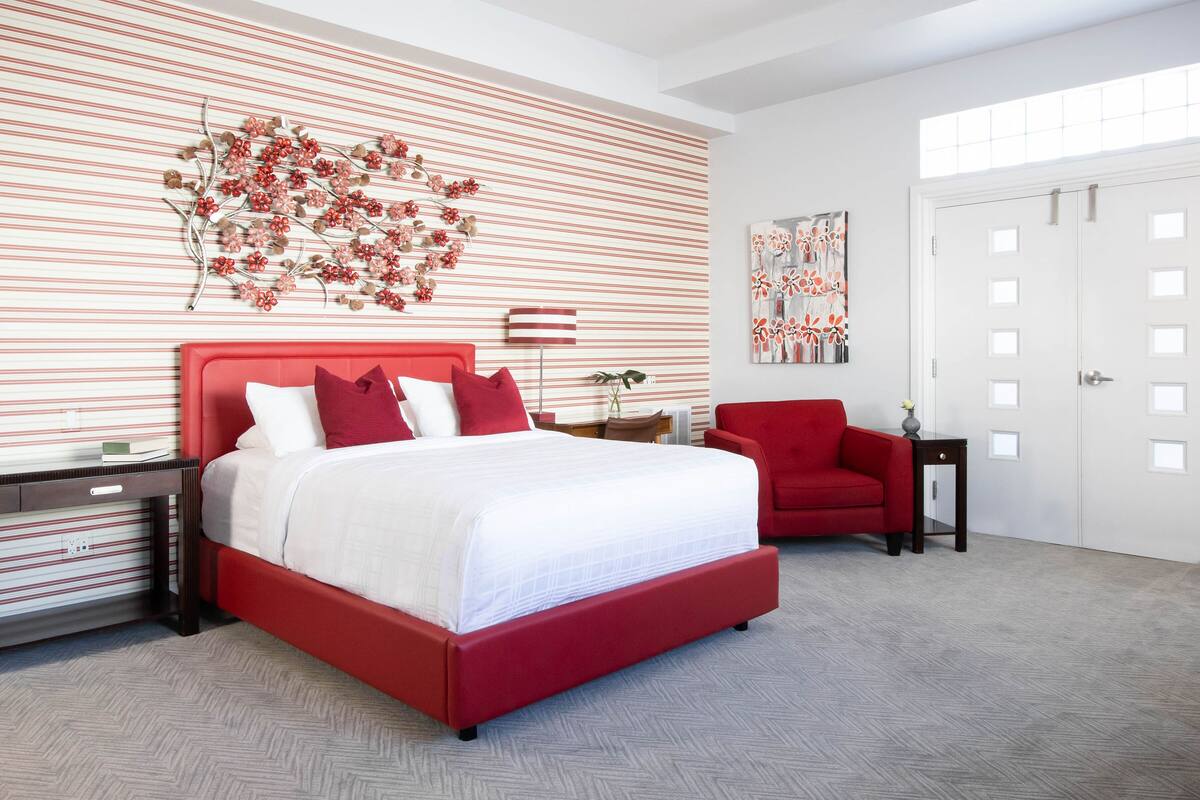 A modern bedroom with a red bed, white walls, and a striped pattern on the wall.