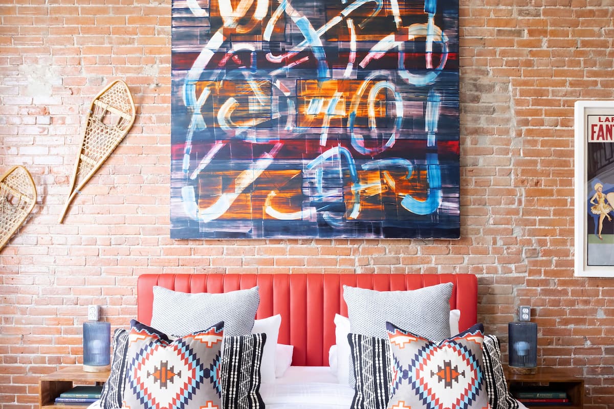 A hotel room with a brick wall, a large abstract painting, a red headboard, and patterned pillows.