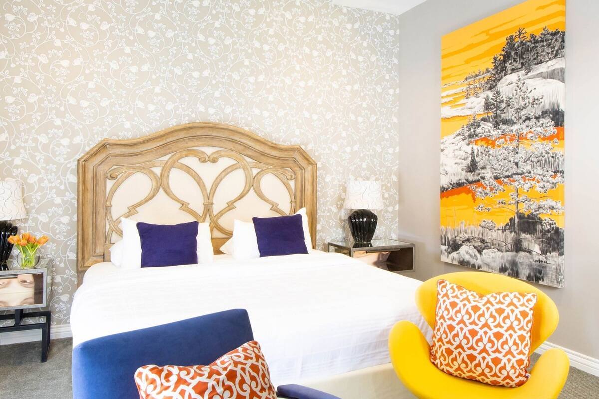 A bedroom with a bed, a nightstand, a lamp, a chair, and a painting on the wall.