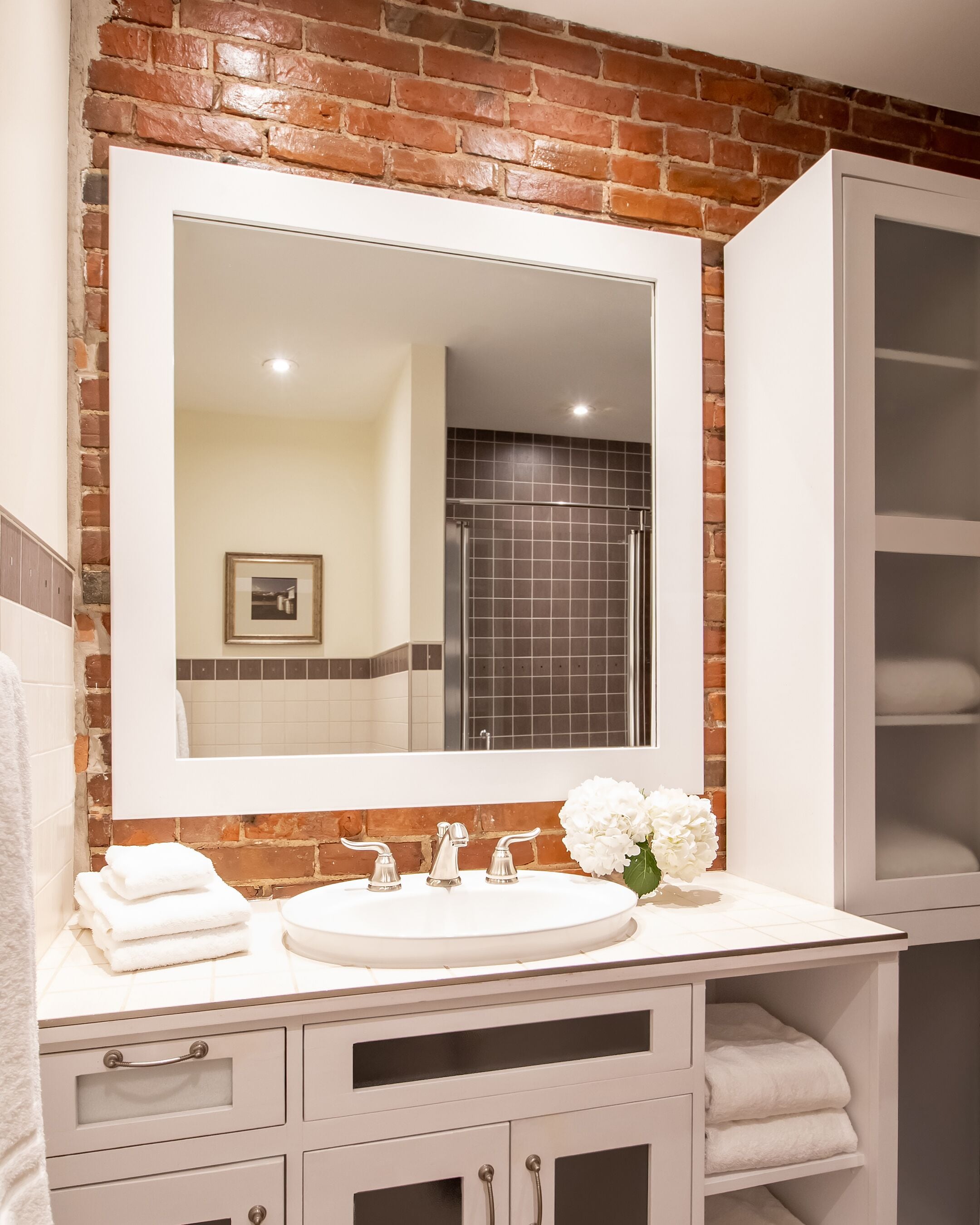 Une salle de bain avec un lavabo blanc, un grand miroir et un mur en briques. Des serviettes sont pliées sur le comptoir, et un vase avec des fleurs y est posé.
