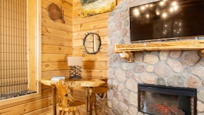 Un interior de cabaña acogedor, con chimenea de piedra, paredes de madera, televisor montado y comedor con mesa y sillas.