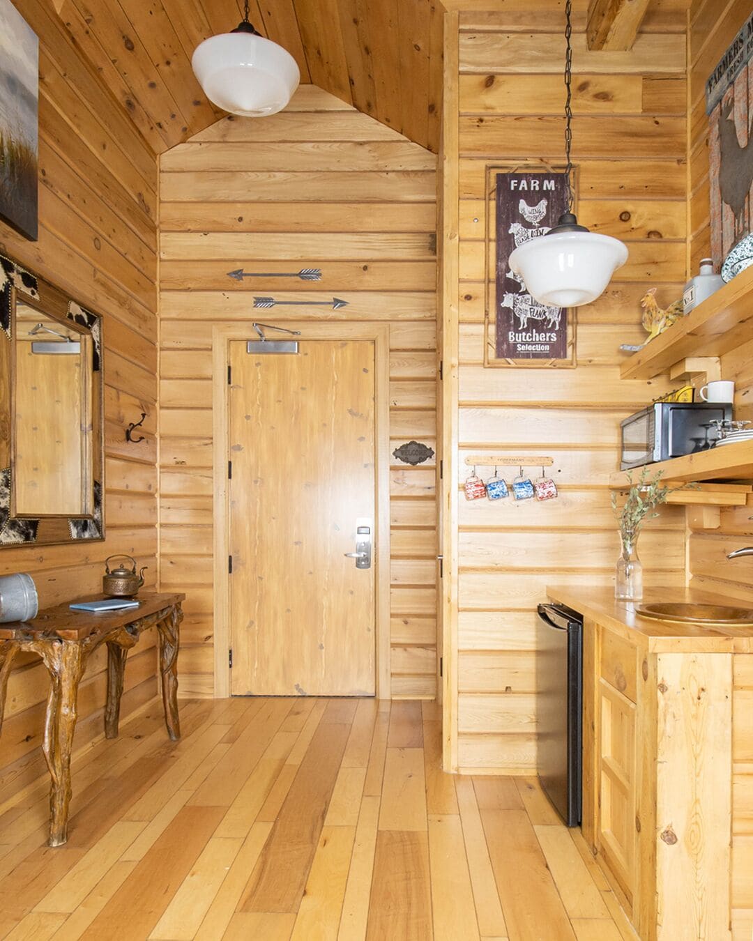 A wooden cabin interior with a wooden door, a table with a kettle, a hanging light fixture, a sign reading "Farm Butchers Selection", and a sink with a mirror above it.