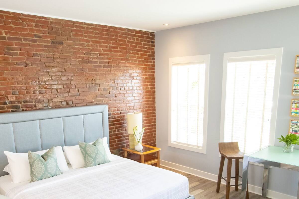 A modern bedroom with a brick accent wall, a bed with a grey headboard, a bedside table with a lamp, and a small desk area with a chair.