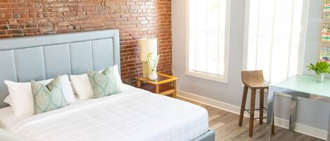 Un dormitorio moderno con una pared de ladrillo decorativa, una cama con cabecera gris, una mesita de noche con una lámpara y una pequeña zona de escritorio con una silla.