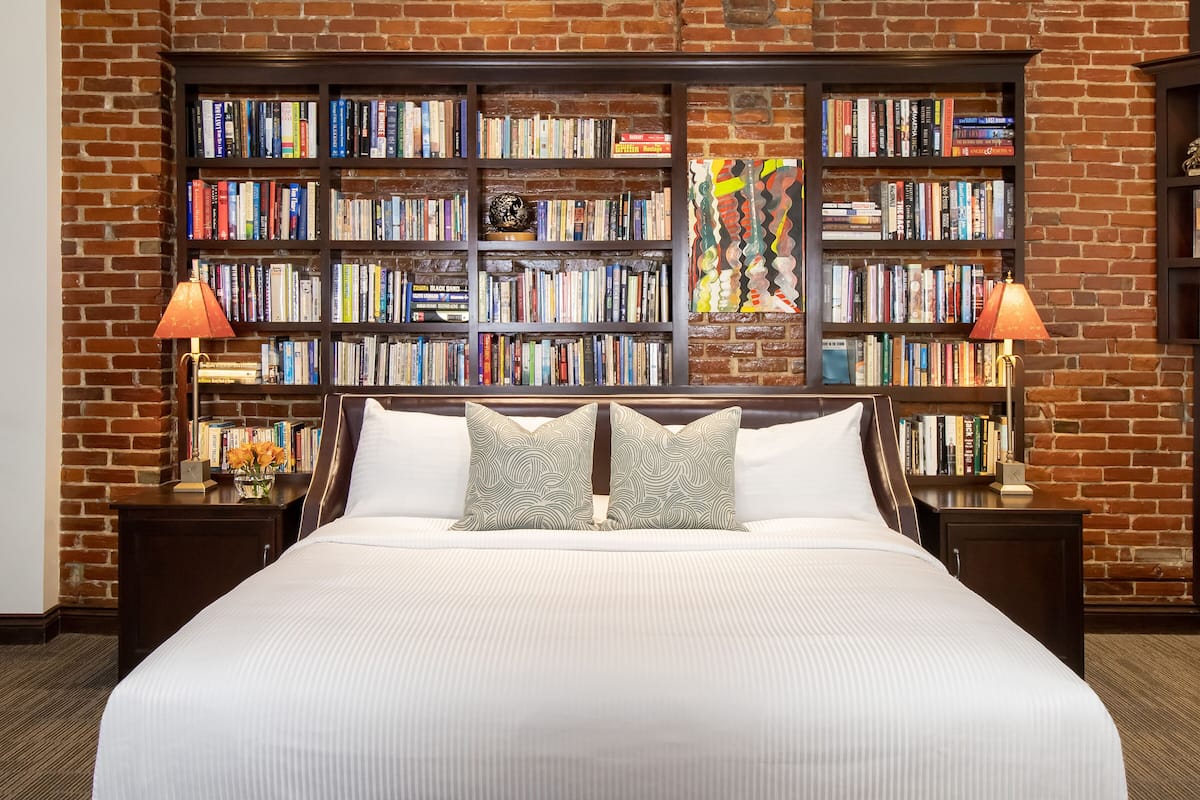 A hotel room with a large bed, two nightstands, a bookshelf, and a brick wall.