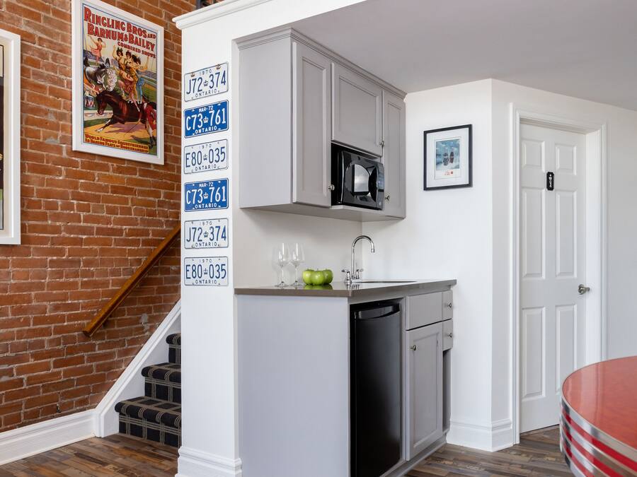 Una cocina compacta con muebles blancos, un microondas y un lavamanos. Una escalera conduce a un área tipo loft con barandilla roja.