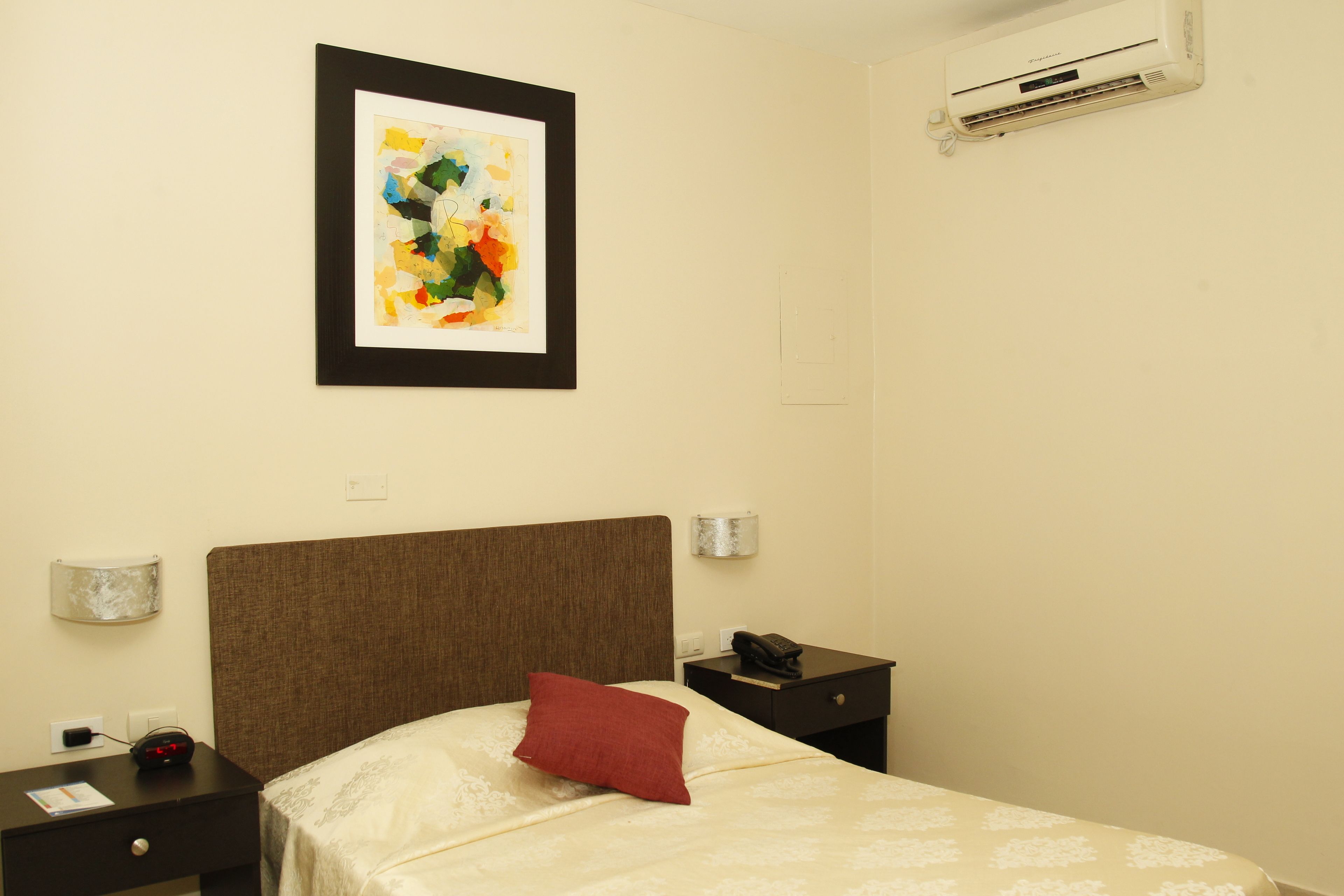 A hotel room with a bed, a nightstand, a framed abstract painting, an air conditioner, and a wall socket.
