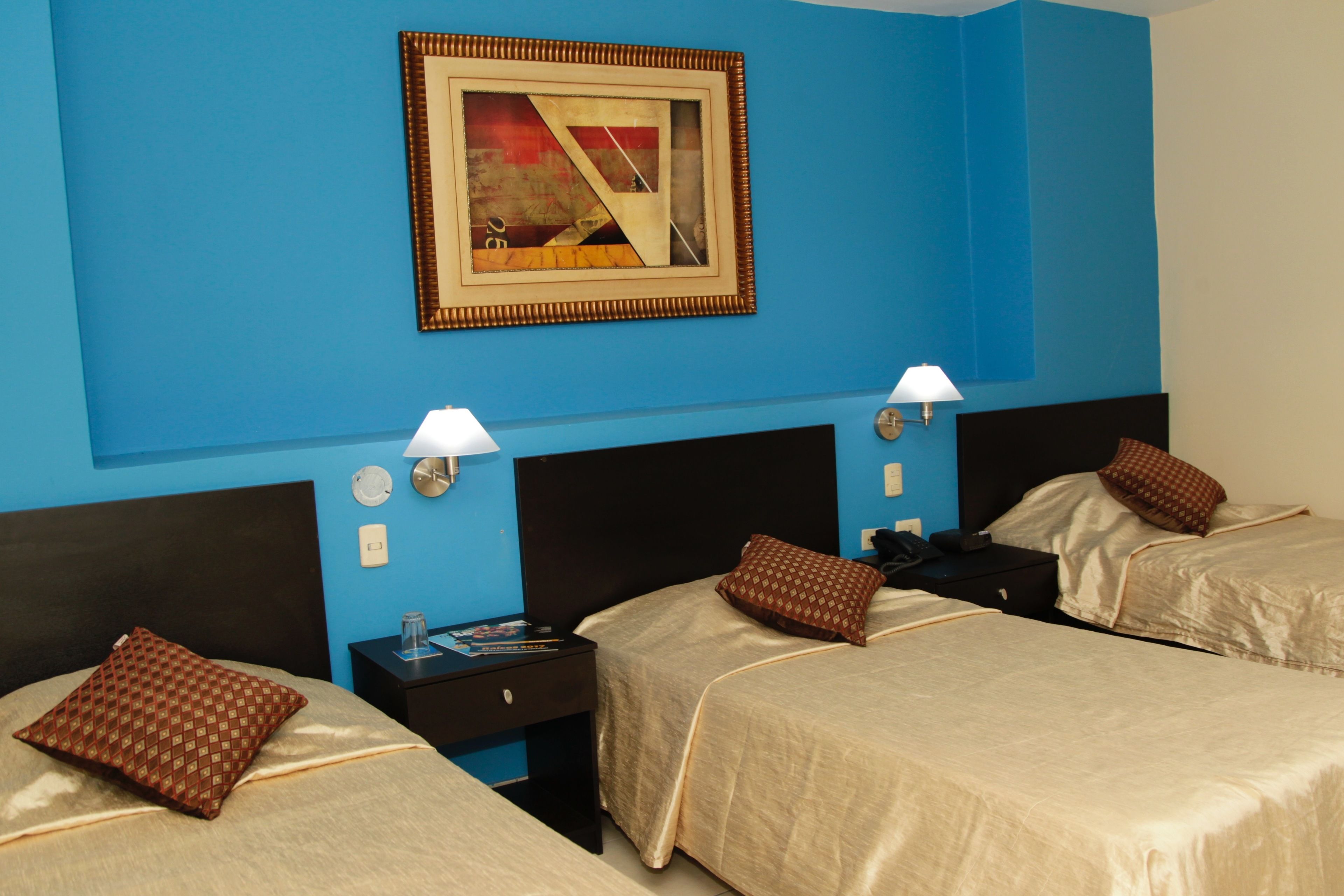 Habitación de hotel con dos camas, cada una con almohada y mesita de noche. Se observa un cuadro en la pared y una lámpara sobre cada mesita.