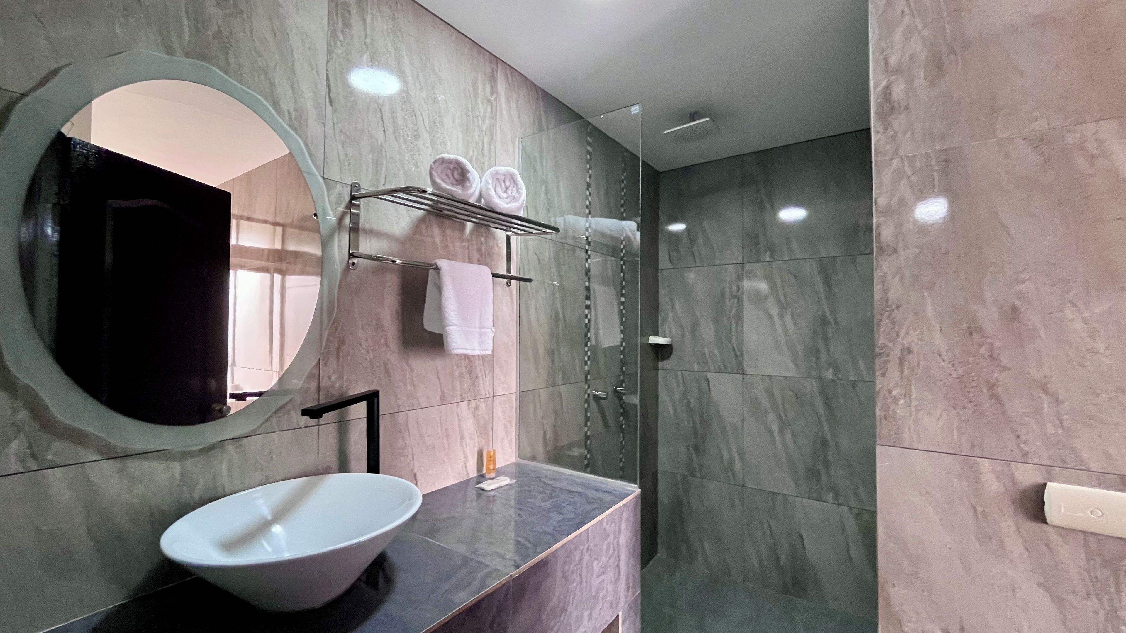 Un baño moderno con un espejo ovalado grande, un lavabo blanco tipo recipiente, un grifo negro y una cabina de ducha de vidrio.