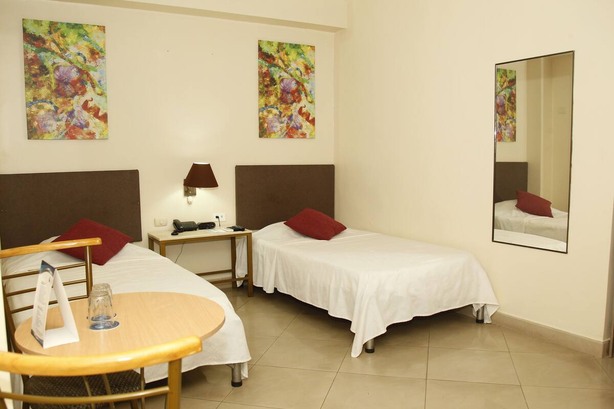 Une chambre d’hôtel avec deux lits, un bureau, une chaise, une table et deux tableaux abstraits accrochés aux murs.