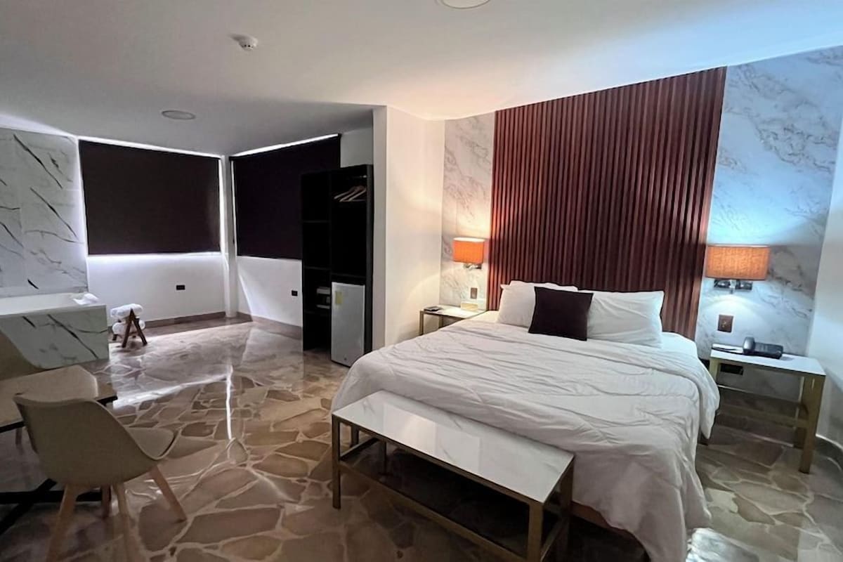 Una habitación de hotel moderna con una cama grande, un escritorio y una silla. Hay dos mesitas de noche con lámparas.