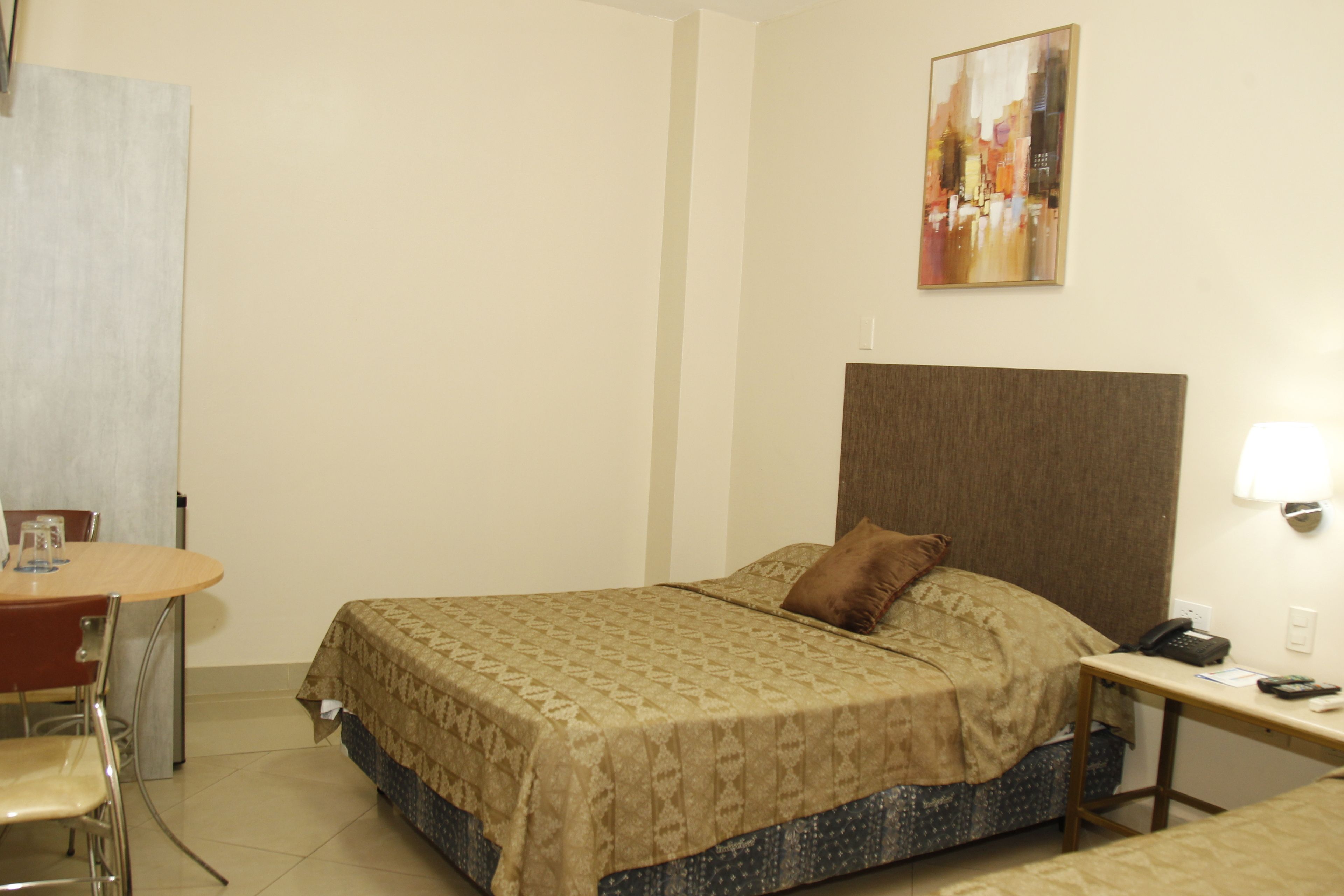 A hotel room with a bed, a desk with a phone, a chair, and a painting on the wall.