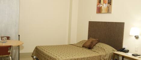 A hotel room with a bed, a desk with a phone, a chair, and a painting on the wall.