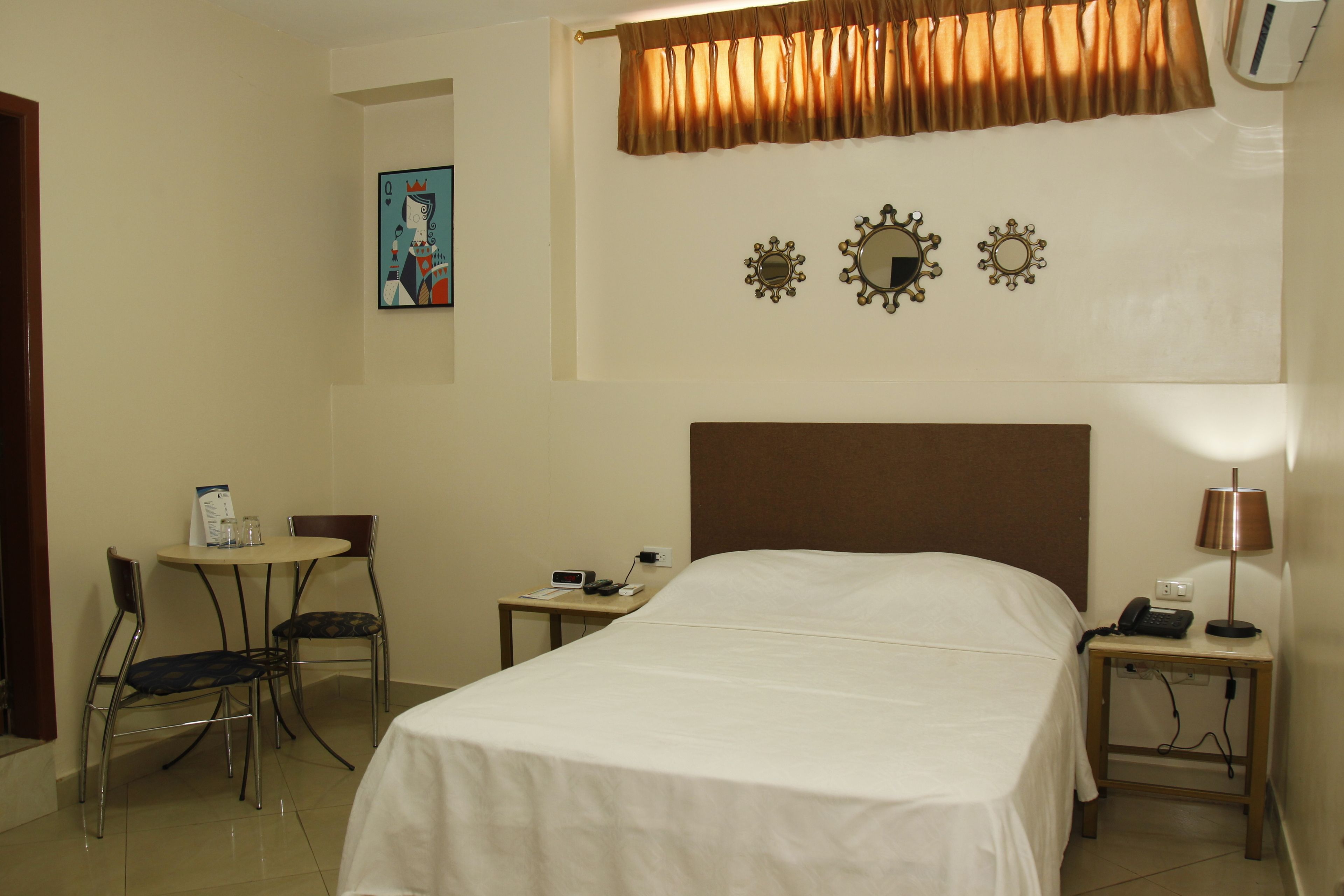Ein Hotelzimmer mit einem Einzelbett, einem kleinen Tisch mit Stuhl, einem Nachttisch mit Lampe und einem an der Wand befestigten Spiegel.