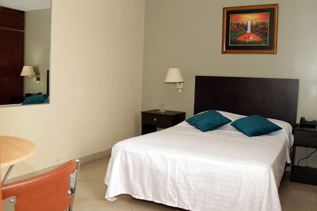 Une chambre d’hôtel avec un lit, un bureau, une chaise, une table de chevet et un tableau au mur.