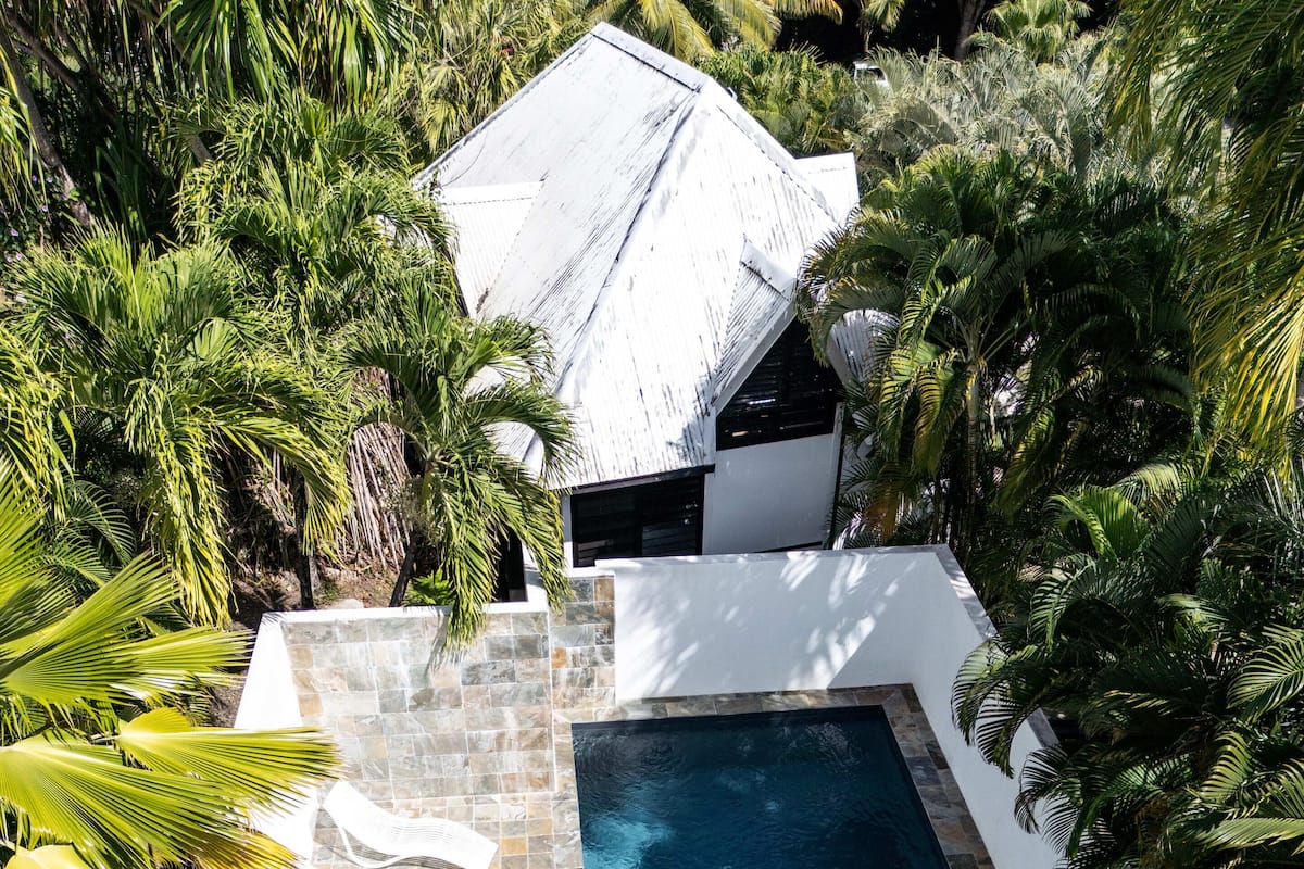 Una piscina rectangular con azulejos de piedra y una estructura de techo blanca, rodeada de abundante follaje.