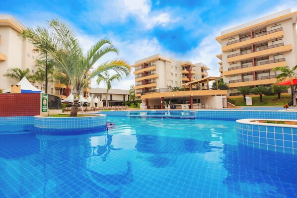 A swimming pool surrounded by multi-story buildings with balconies, palm trees, and outdoor seating areas.