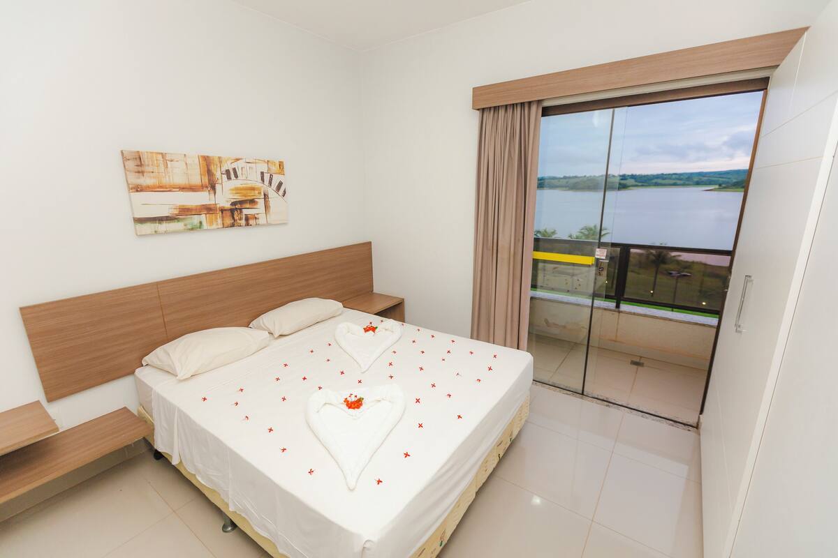 A bedroom with a bed, a nightstand, a painting on the wall, and a view of a body of water through a sliding glass door.