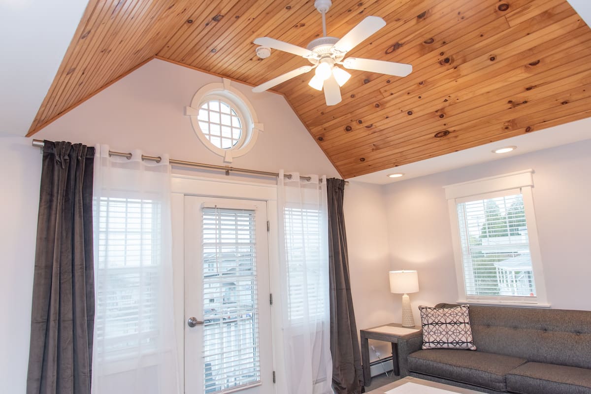 A room with a wooden ceiling, a ceiling fan, a window with blinds, a sofa with a patterned cushion, and a lamp on a side table.