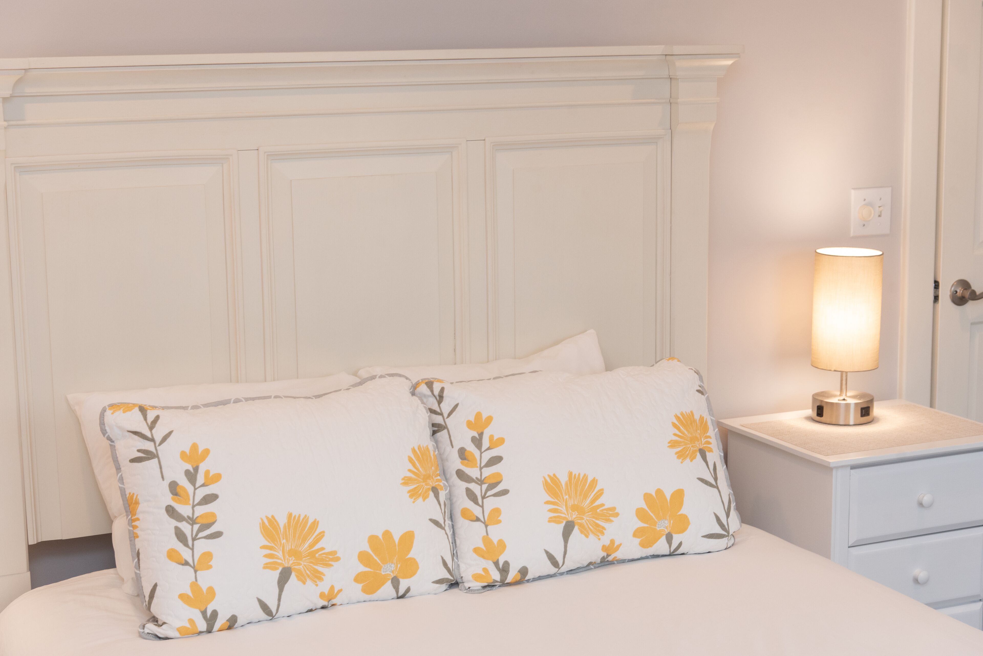 A neatly made bed with two pillows featuring a floral pattern, a bedside table with a lamp, and a white nightstand.