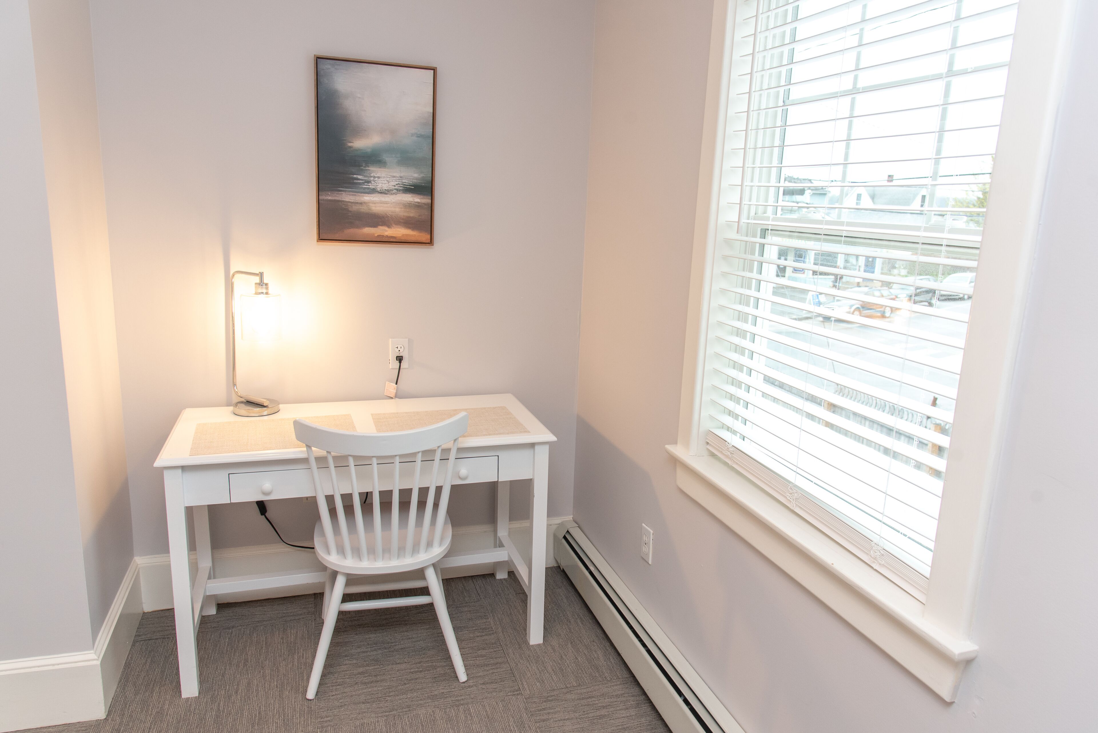 A white desk with a chair, a wall-mounted lamp, and a painting above it.